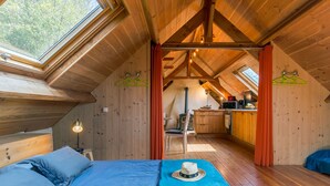 Un dormitorio acogedor en el ático con una cama, una manta azul y un sombrero sobre la cama. Hay una kitchenette con estufa y microondas.