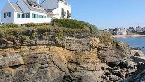 Un edificio bianco con persiane verdi, arroccato su una scogliera a picco su una spiaggia.