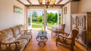 Ein Zimmer mit Sofa, Sesseln, Tisch und Schrank. Eine Tür führt in einen Garten.