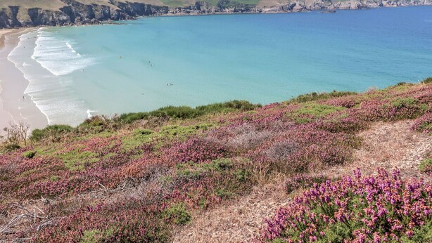 Paisaje costero con playa, acantilados y un faro a lo lejos.