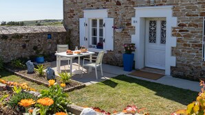Una casa de piedra con una puerta blanca, un pequeño jardín con flores y una mesa dispuesta para dos.