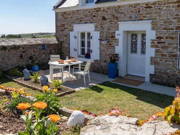 A stone house with a white door, a small garden with flowers, and a table set for two.