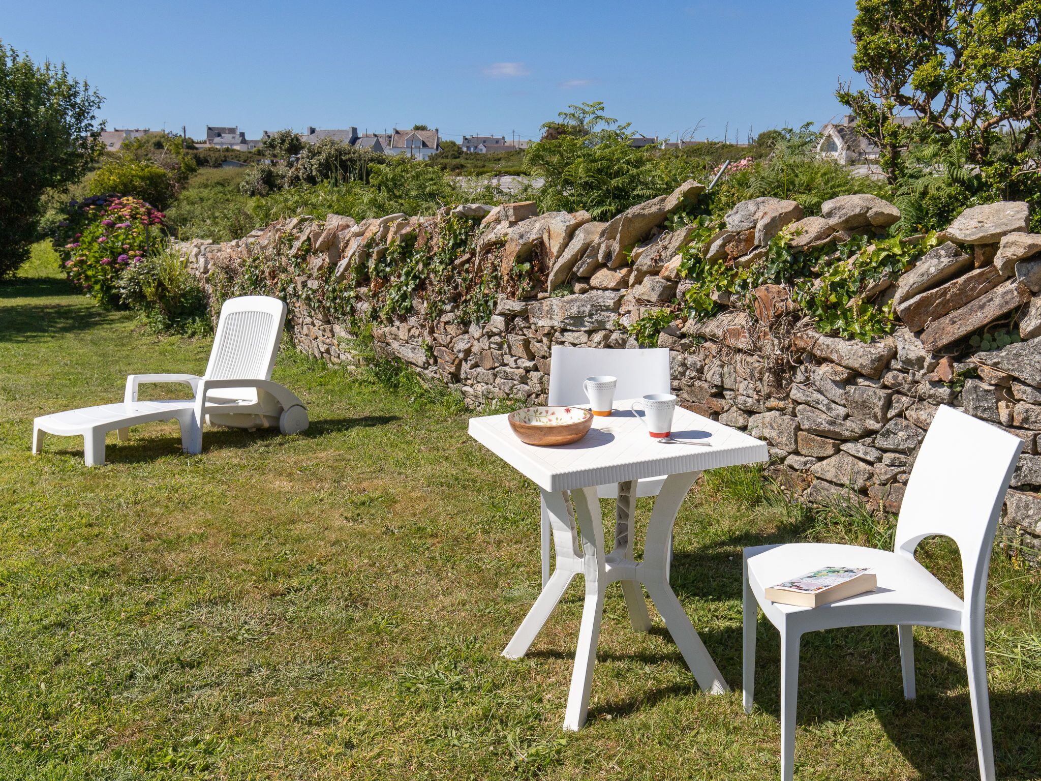 An outdoor seating area with white plastic chairs and a table, surrounded by a stone wall and greenery.
