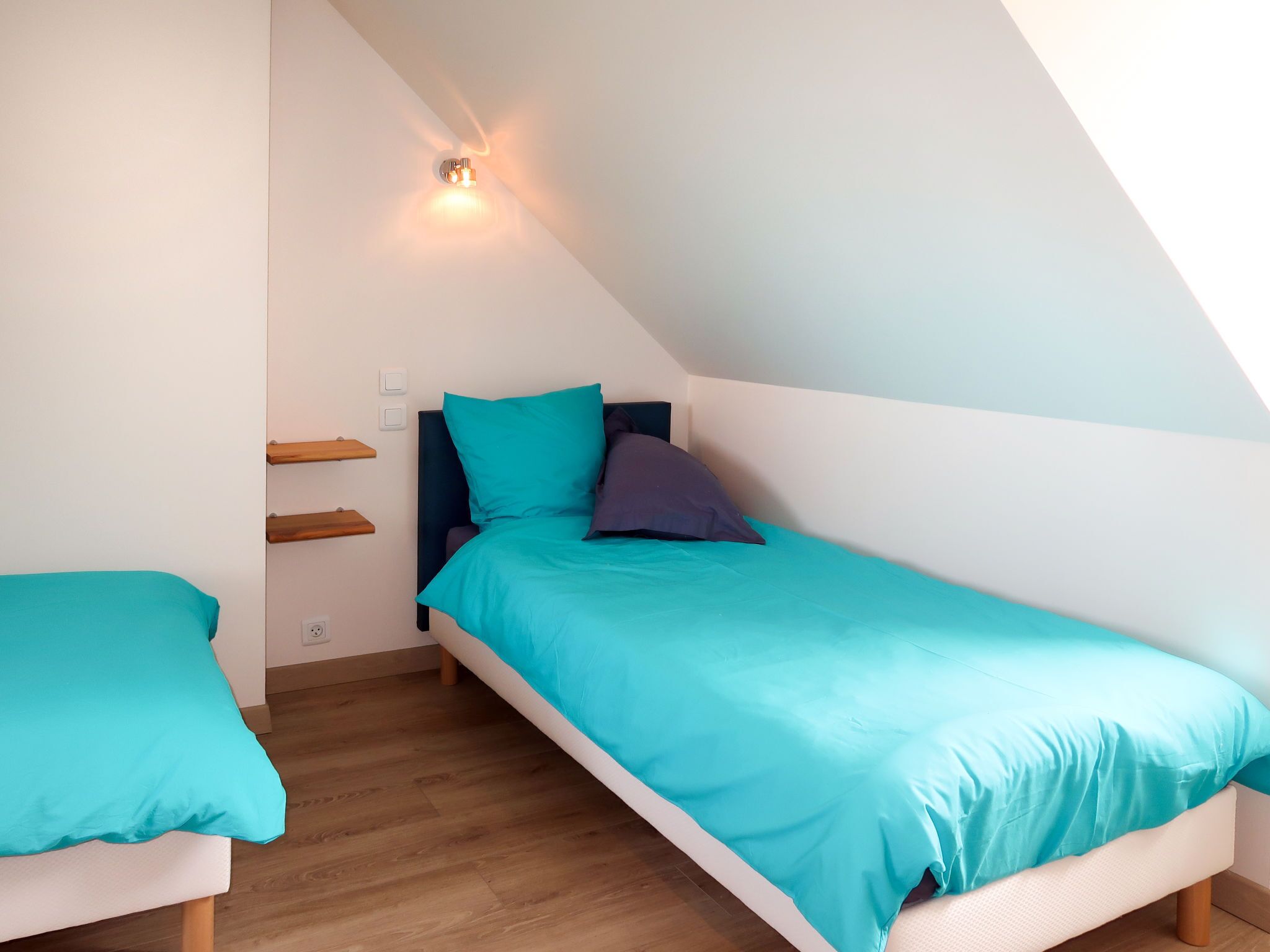 A small, triangular room with a bed and a smaller bed against the wall, a light fixture above the larger bed, and a wooden floor.
