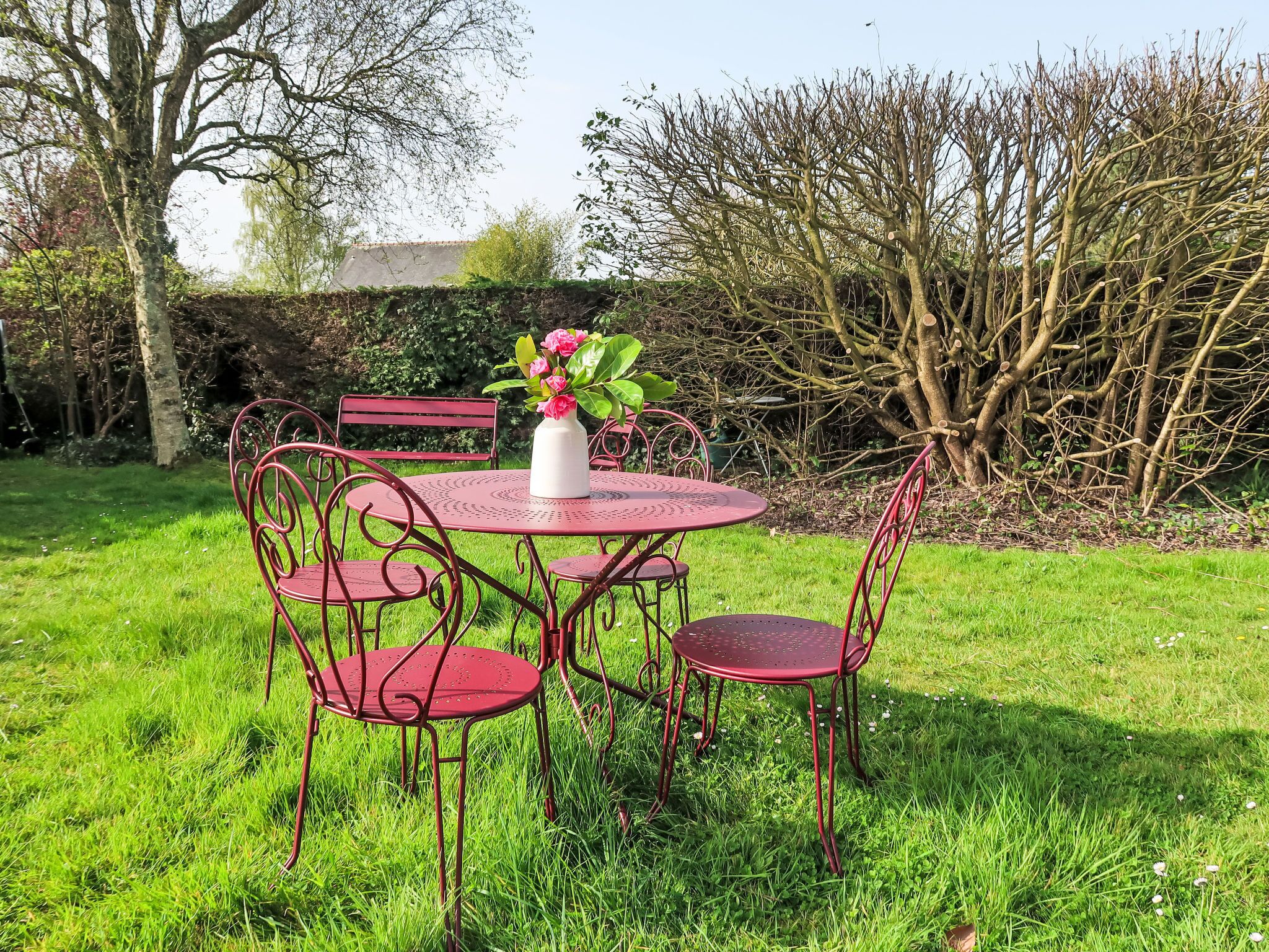 A round metal table with four red chairs, a white vase with pink flowers, and a hedge in the background.