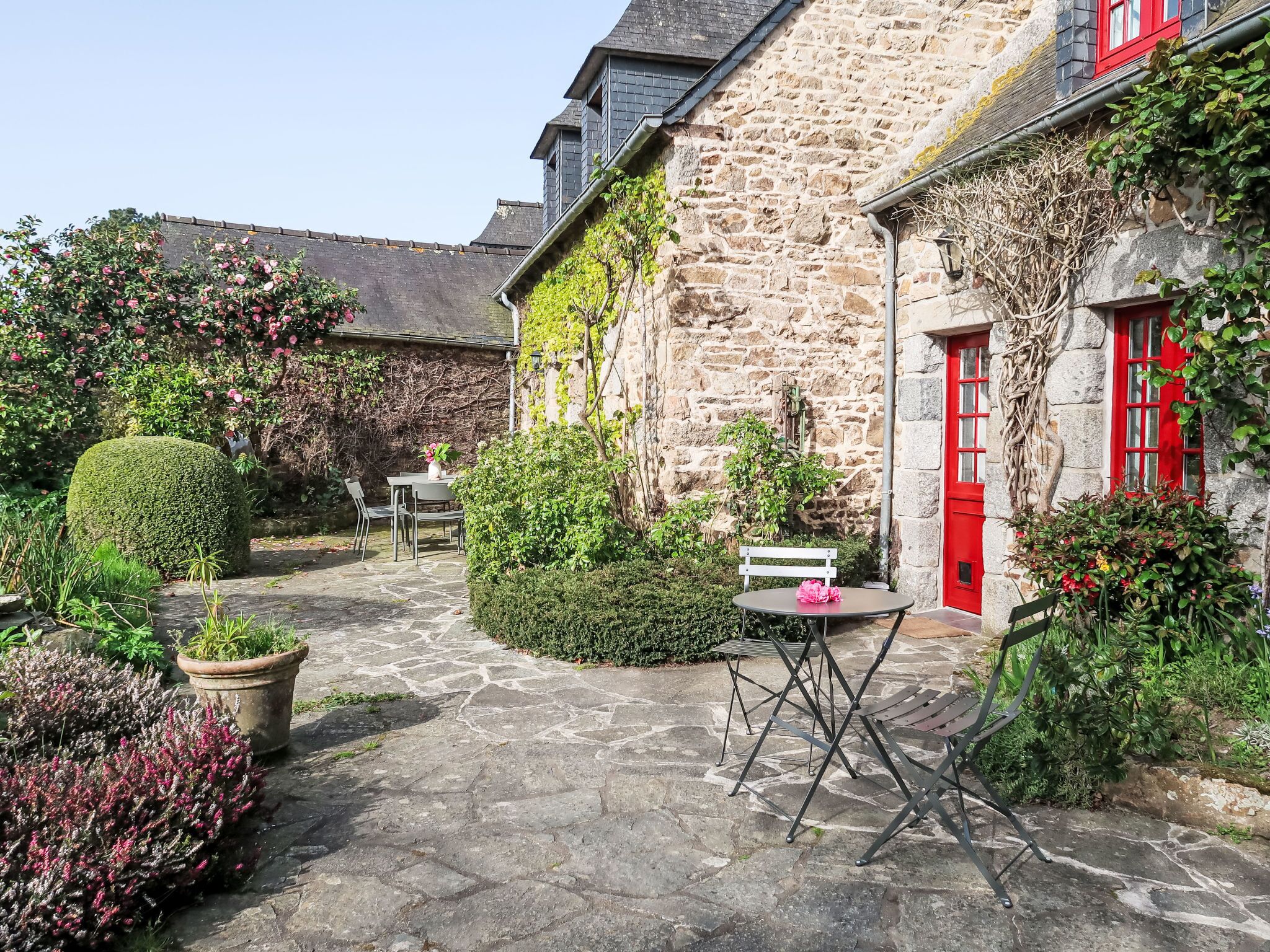 A stone building with a red door and window shutters, surrounded by a stone-paved courtyard with a table and chairs, and various plants.