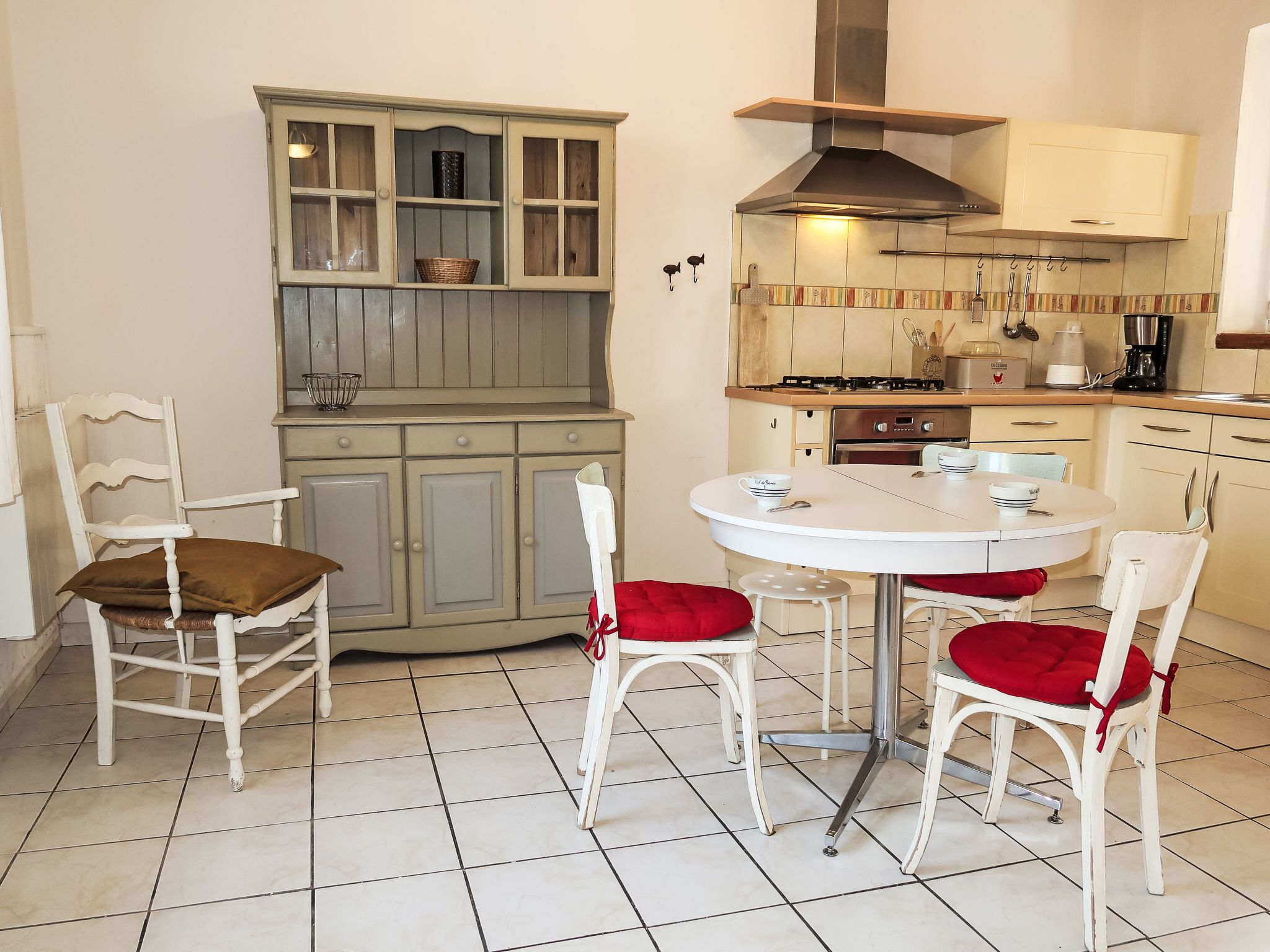 A compact kitchen with a round dining table, white chairs with red cushions, and a cabinet with glass doors.