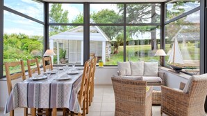 A dining area with a table set for six, surrounded by wicker chairs, large windows offering a view of a garden and a small sailboat model. - Chante-Brise by Interhome (Plérin)