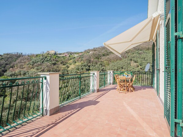 A balcony with a table and chairs, a white umbrella, and a view of a hillside.