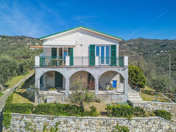A two-story house with a balcony and arches, surrounded by greenery and hills.