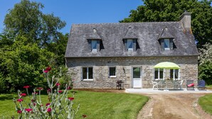 Une maison en pierre avec une allée pavée, une terrasse dotée de mobilier d’extérieur et un jardin agrémenté de fleurs rouges.
