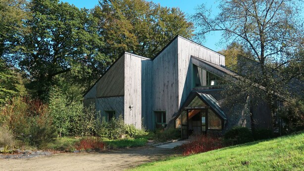 Une maison moderne en bois, dotée d’un toit en pente et de grandes fenêtres, entourée d’arbres et d’une zone herbeuse.