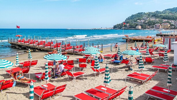 A beach with red and white striped umbrellas and chairs, a pier extending into the sea, and buildings in the background.