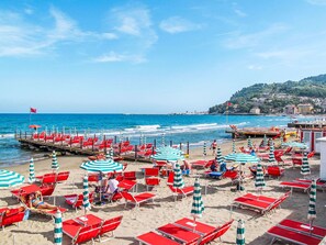 Une plage avec des parasols et des chaises à rayures rouges et blanches, une jetée qui s’avance dans la mer et des bâtiments en arrière-plan.
