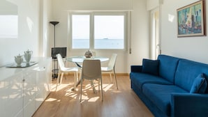A modern living room with a blue sofa, a glass dining table with chairs, a TV, and a view of the sea through large windows.