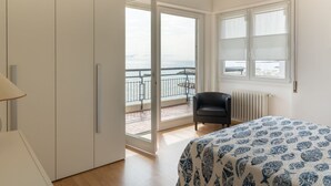 A bedroom with a bed, a chair, a radiator, and a view of the sea through a balcony door.