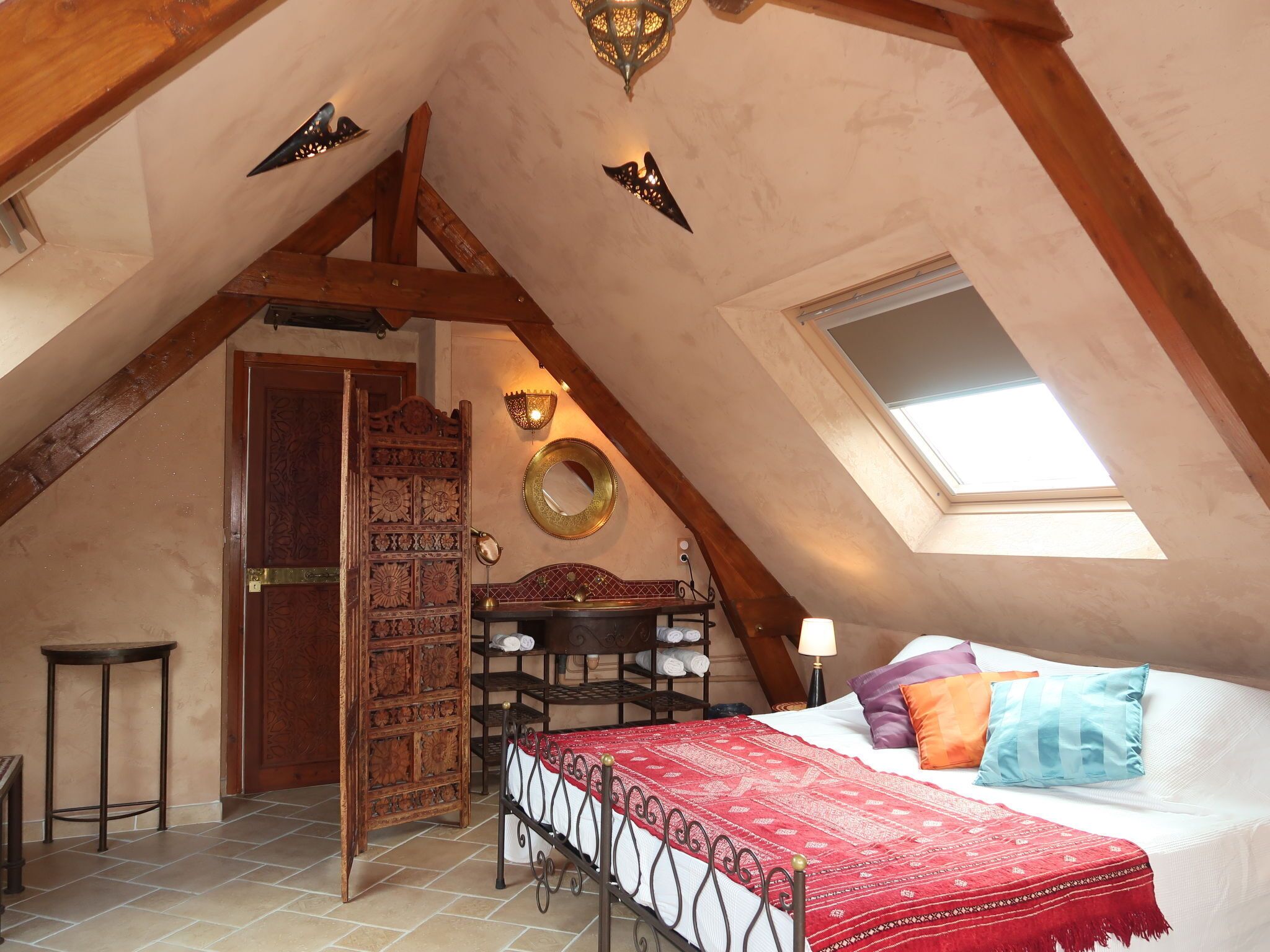 A cozy attic bedroom with a sloped ceiling, a wooden wardrobe, a bed with a red patterned bedspread, a round mirror, and a skylight.
