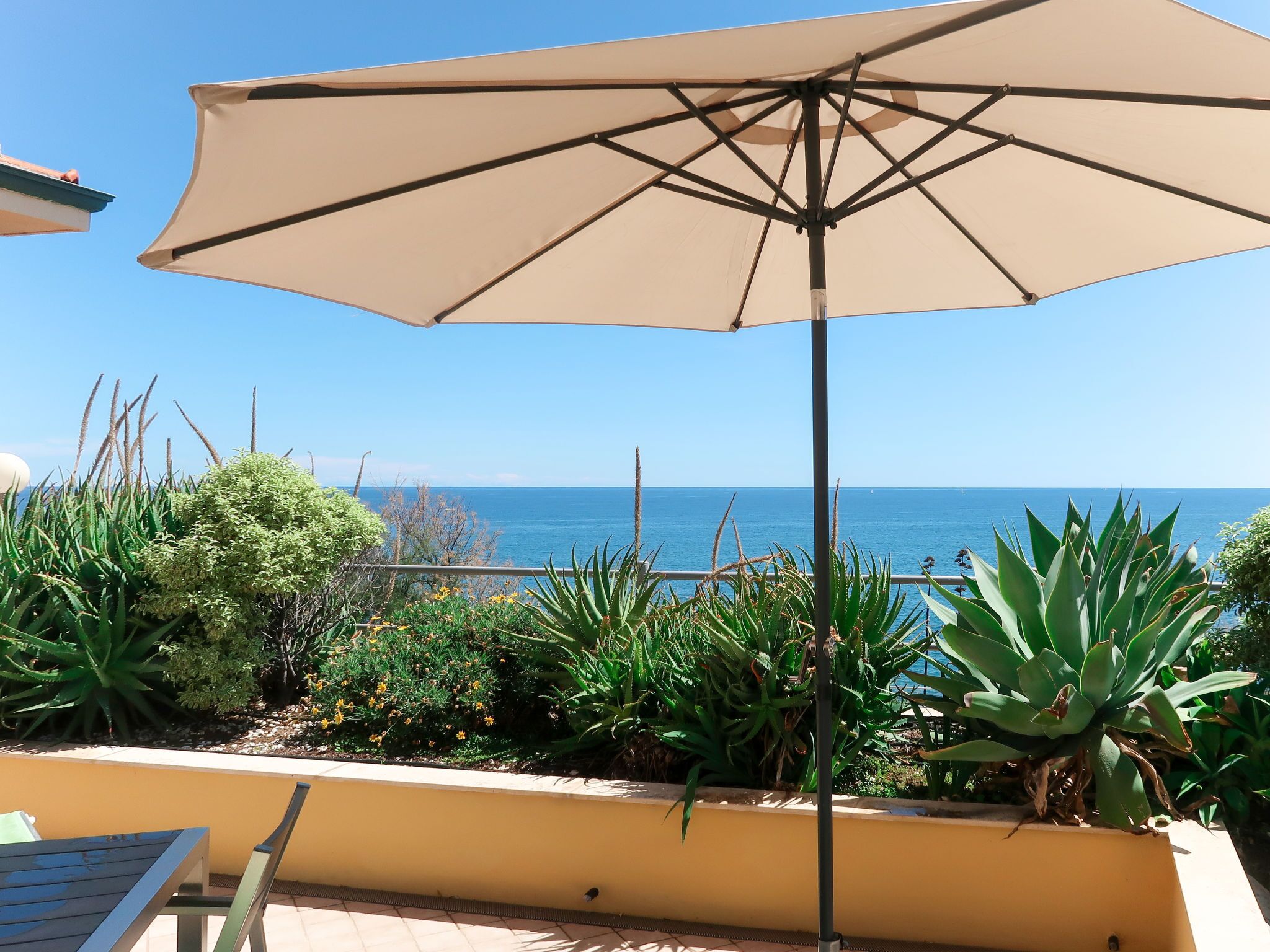 A patio with a beige umbrella, green plants, and a view of the ocean.