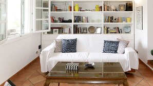 Una sala de estar con un sofá blanco, una mesa de centro de vidrio y una estantería llena de libros y objetos decorativos.