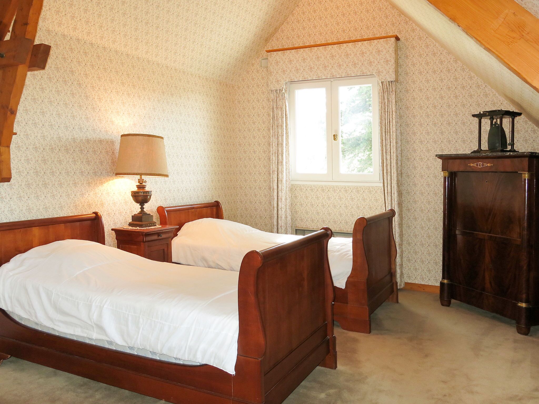 A bedroom with two wooden beds, a nightstand with a lamp, a window with curtains, and a wooden wardrobe.