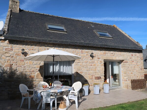 A stone building with a tiled roof, a patio with a table and chairs, and a large umbrella.