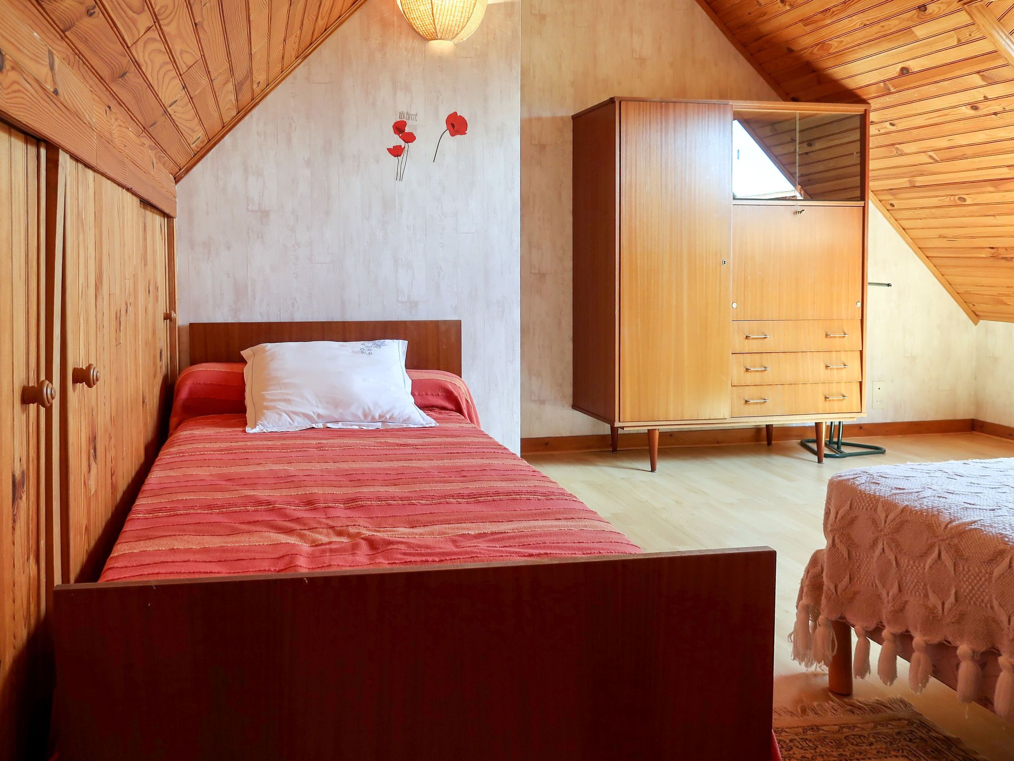 Quarto com teto de madeira, cama com colcha vermelha, guarda-roupa de madeira e uma pequena mesa com toalha branca.