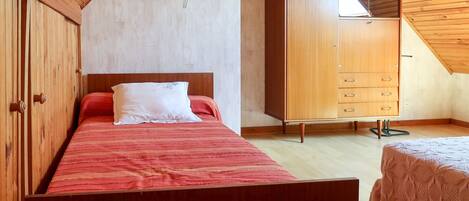 Quarto com teto de madeira, cama com colcha vermelha, guarda-roupa de madeira e uma pequena mesa com toalha branca.