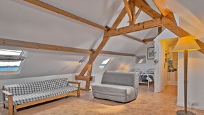 Ein gemütliches Schlafzimmer im Dachgeschoss mit Holzbalkendecke, einem karierten Sofa, einem grauen Sessel und einer Stehlampe.