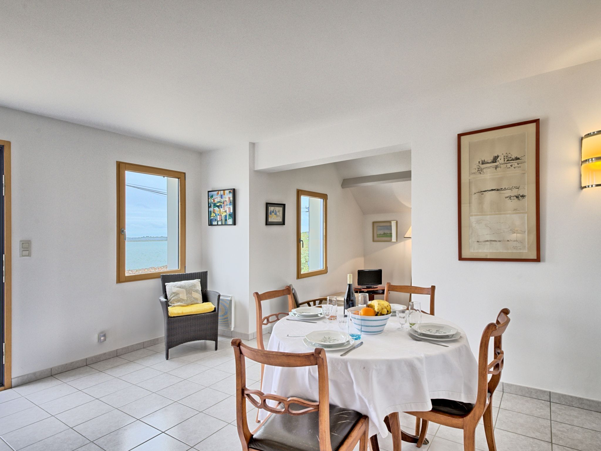 Un comedor con una mesa redonda dispuesta para cuatro personas, rodeada de sillas de madera. Hay una ventana con vista al mar, y las paredes están decoradas con obras de arte enmarcadas.