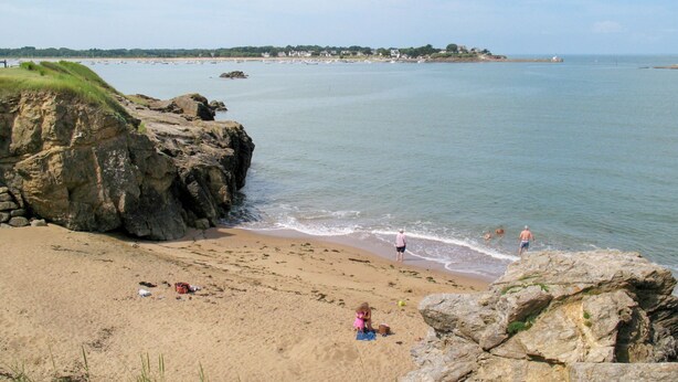 Ein Strand mit Felsen, ruhigem Wasser und einer fernen Küste mit Gebäuden.