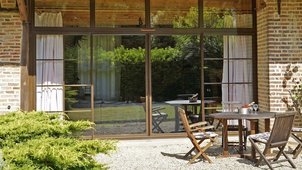 A patio area with a table and chairs, a wooden pergola, and a view of a garden through large sliding glass doors. - Alexandre by Interhome (La Chapelle-Saint-Sauveur)