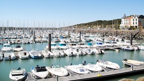 A marina with numerous boats docked, a building with a red roof, and a rocky hillside in the background. - Les Oliviers by Interhome (Talmont Saint Hilaire)