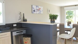Una cocina moderna con una isla central, lavavajillas, contenedor de basura, zona de comedor con sillas y una planta en maceta.