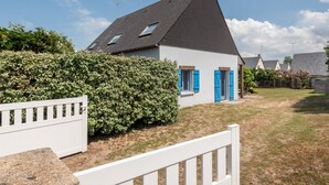 A house with a dark roof, white walls, and blue shutters. There is a white fence in the foreground and a hedge behind the house.