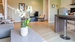 Un salon moderne avec un canapé gris, une table basse blanche agrémentée d’une plante, un téléviseur et un coin cuisine avec un tabouret de bar.
