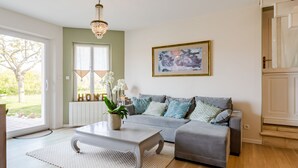Un salon avec un canapé gris, une table basse blanche et un lustre. Il y a un tableau au mur et une porte sur laquelle est apposée une plaque indiquant « MAISON ».