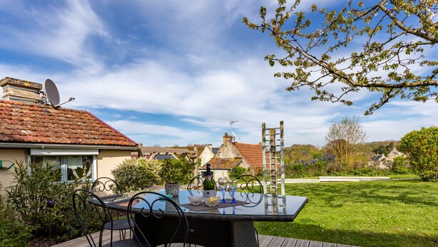 Une terrasse en bois avec une table et des chaises en verre, une bouteille de vin et un saladier de fruits.