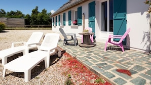 Een gebouw met witte muren en turquoise luiken, een grindpad en buiten zitplaatsen met witte en roze stoelen.