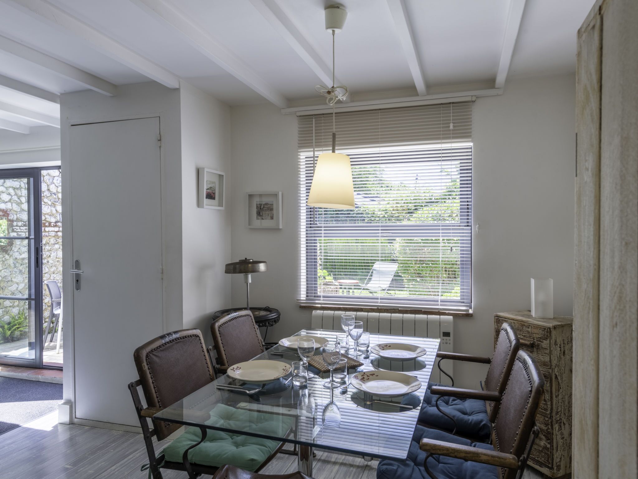 Un coin repas avec une table en verre, des chaises et une fenêtre équipée de stores.