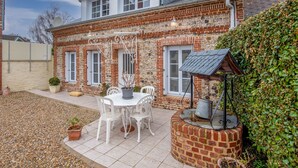 Ein zweistöckiges Haus mit Ziegelaußenwand und weißen Verzierungen. Es gibt eine Terrasse mit Tisch und Stühlen sowie einen kleinen Gartenbereich mit Pflanzen und einem Vogelbad.
