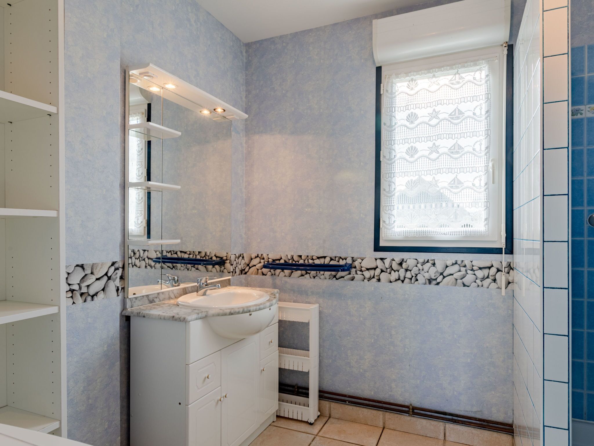 Ein Badezimmer mit Marmorwaschbecken, weißen Schränken, einem Spiegel und einem Fenster mit dekorativer, gemusterter Gardine.