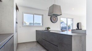 A modern kitchen with a large island, a hanging light fixture, and a view of the outdoors through large windows.