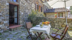 Un bâtiment en pierre au charme rustique, avec une table dressée pour deux personnes, agrémentée d’un panier de fruits et d’une bouteille d’huile d’olive.