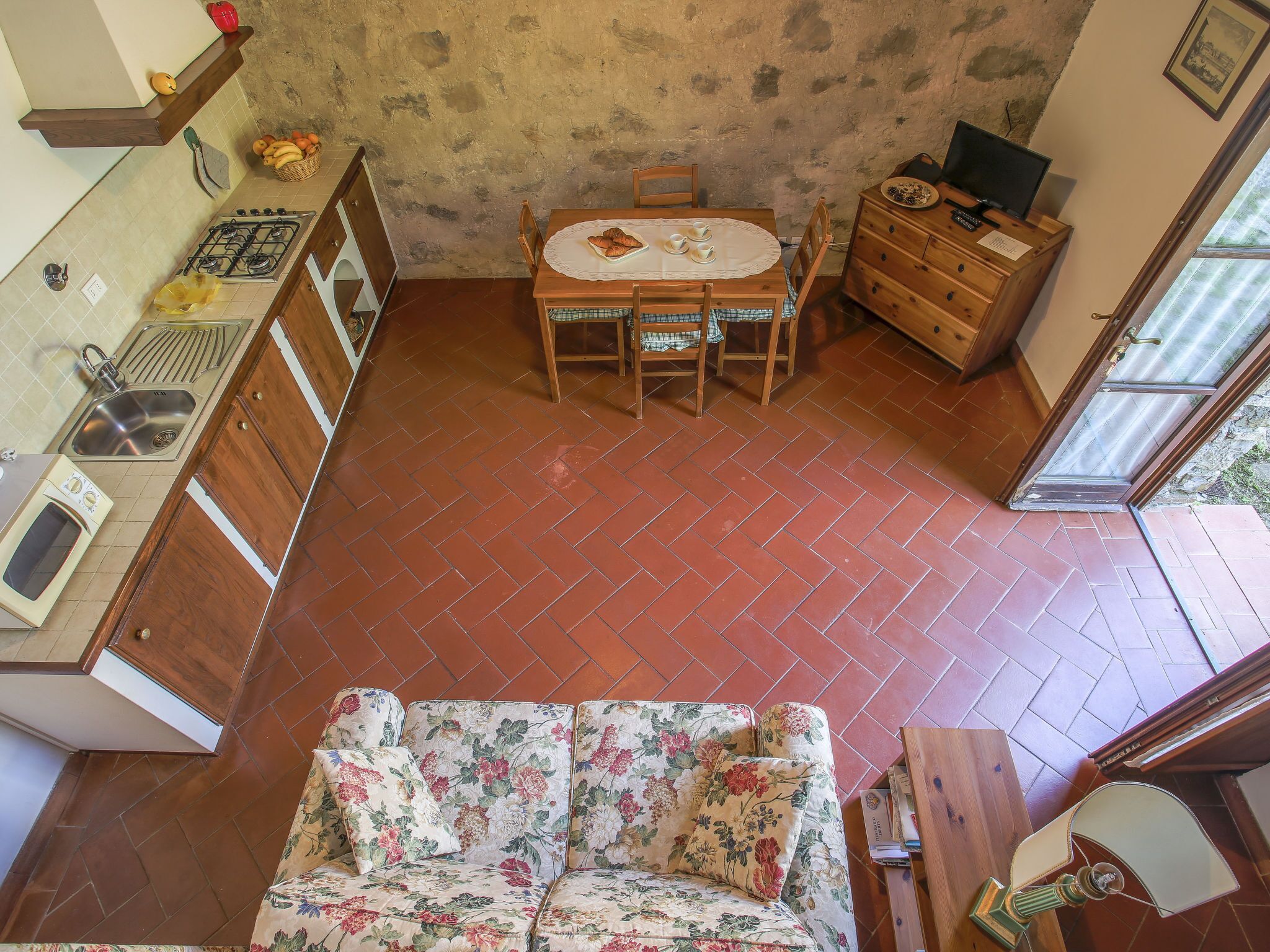 Een keuken met een gasfornuis, een spoelbak en houten kastjes. Er is een eettafel met stoelen en een bloemenbank. Ook zichtbaar is een houten dressoir en een televisie.