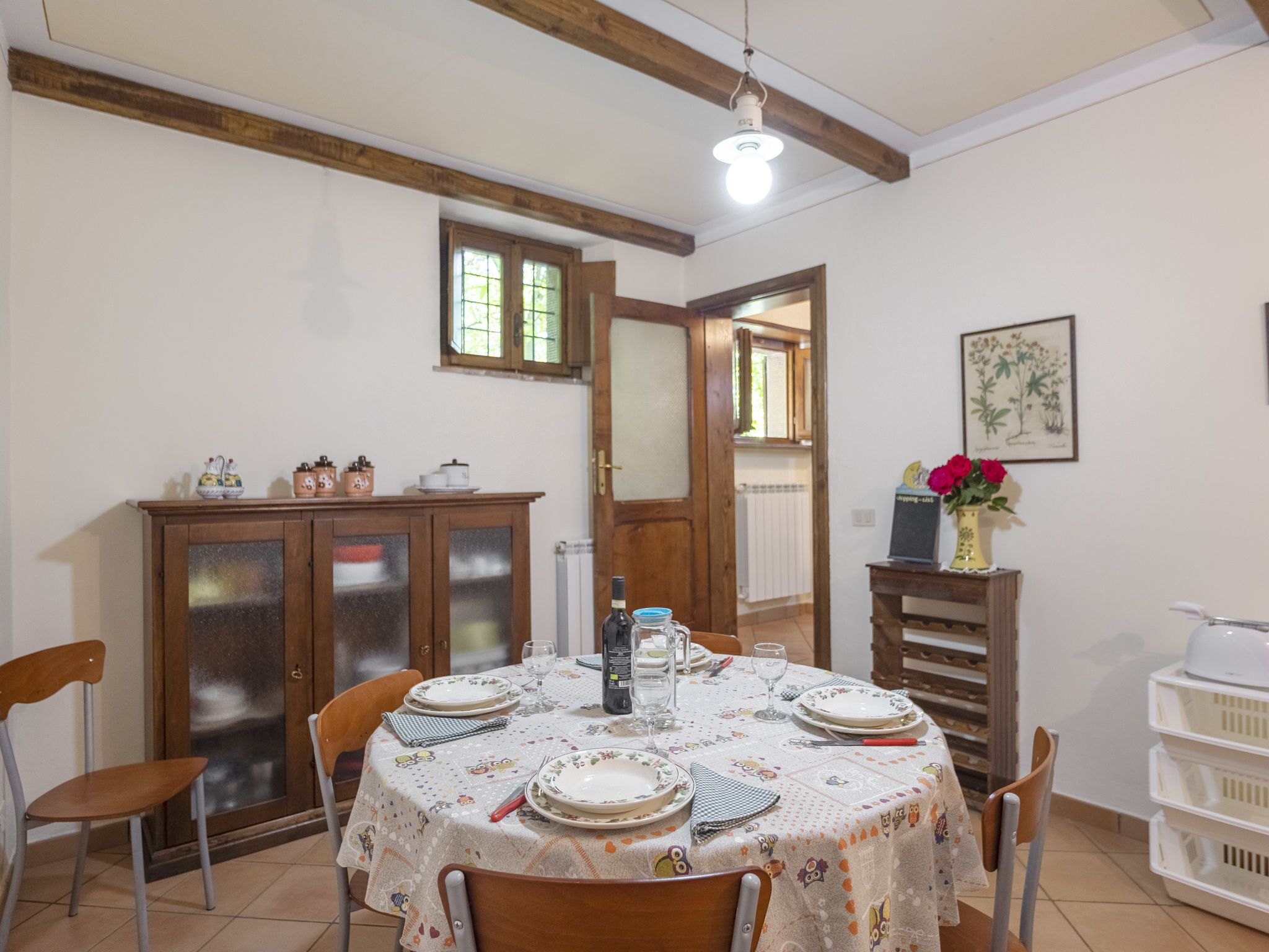 Un comedor con una mesa redonda dispuesta para cuatro personas, rodeada de sillas. Se observa un aparador de madera con puertas de cristal, un radiador e ilustración botánica enmarcada en la pared.