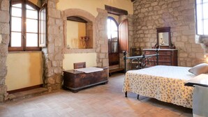 A rustic bedroom with stone walls, wooden beams, a bed with a floral bedspread, a wooden dresser, and a bench.