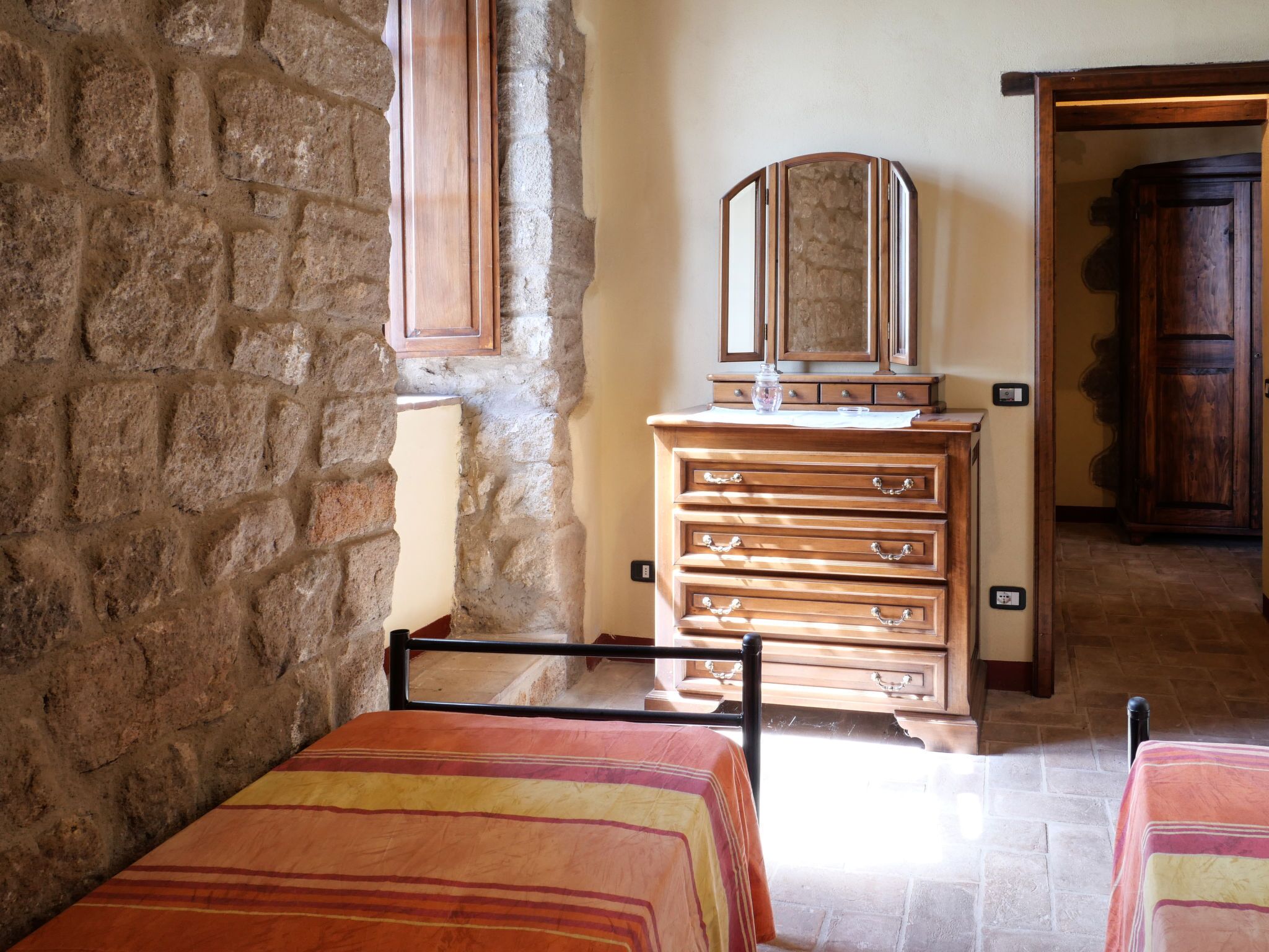 Une chambre avec un mur en pierre, une commode en bois, un miroir et deux lits avec une literie à rayures.