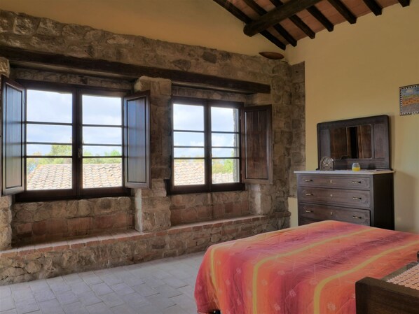 A bedroom with stone walls, a bed with a colorful blanket, a dresser, and a large window with shutters. - Torretta - Borgo la Civitella by Interhome (Roccastrada)