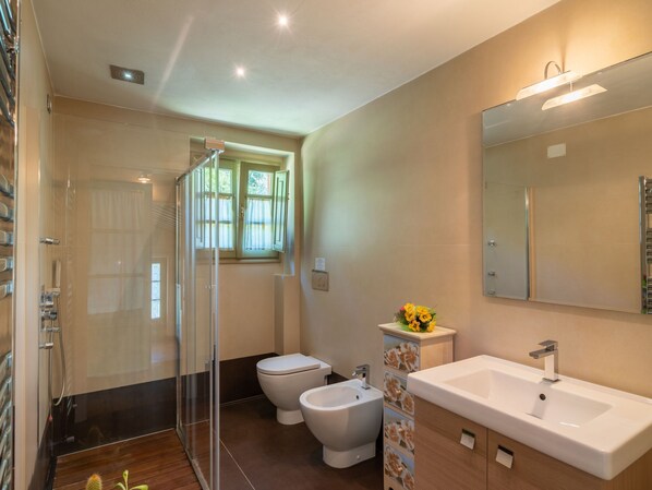 A modern bathroom with a glass shower enclosure, two toilets, a bidet, and a large sink with a mirror.