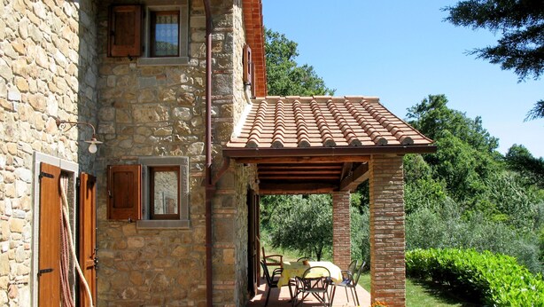A stone building with a tiled roof, a covered patio with outdoor furniture, and a well-maintained garden.